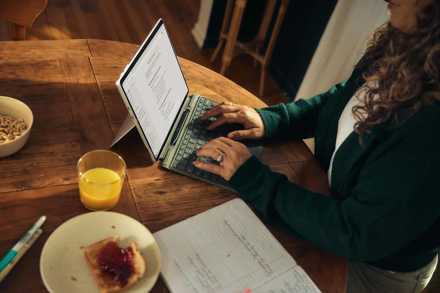 Woman typing on a tablet with a keyboard at a wooden table, next to breakfast and notes