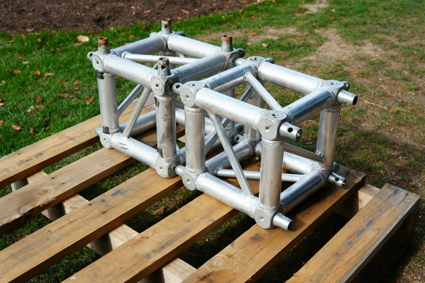 Aluminium truss sections lie on a wooden pallet in the grass