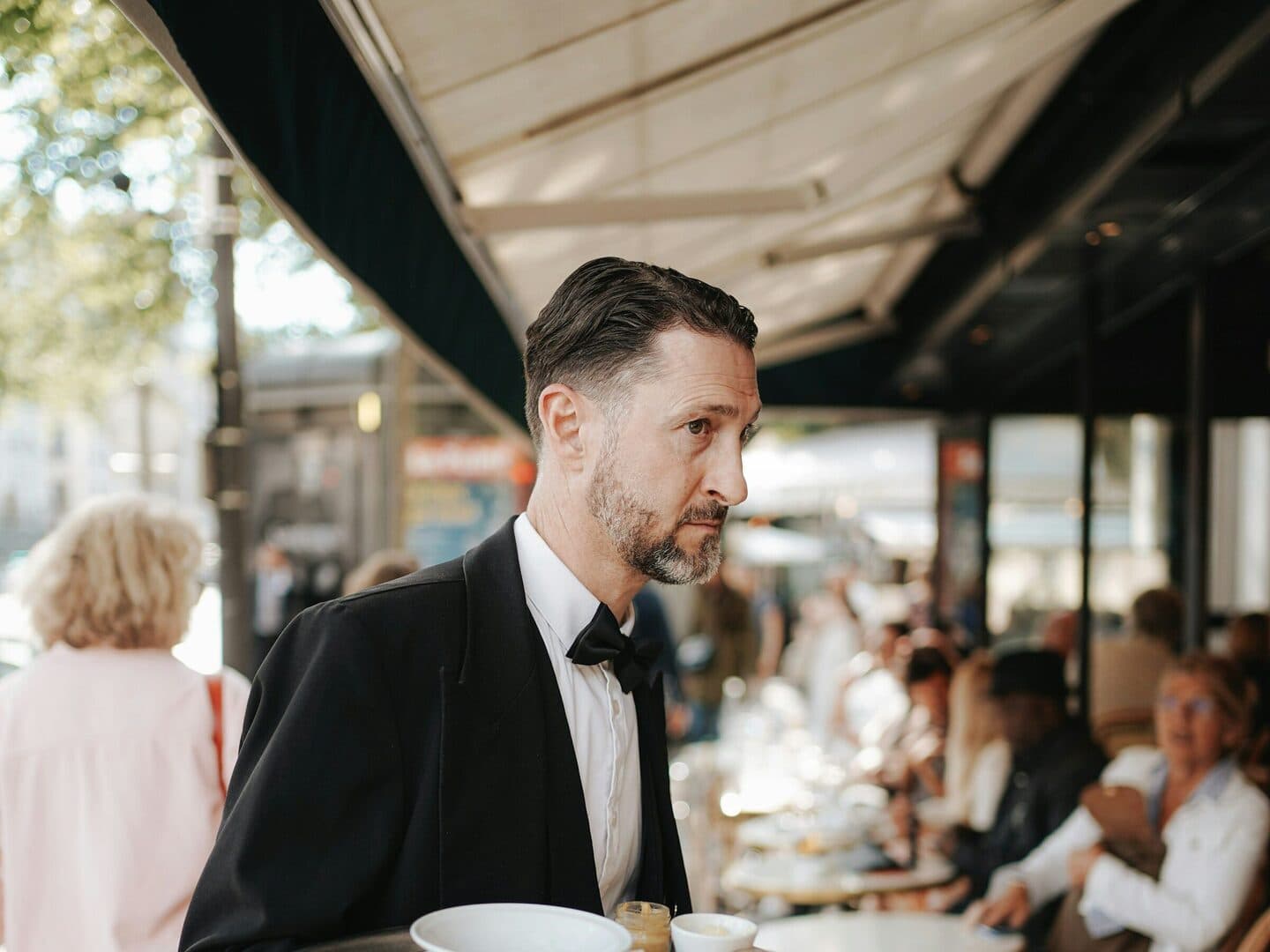 Waiter in a tuxedo carries a tray across a café terrace