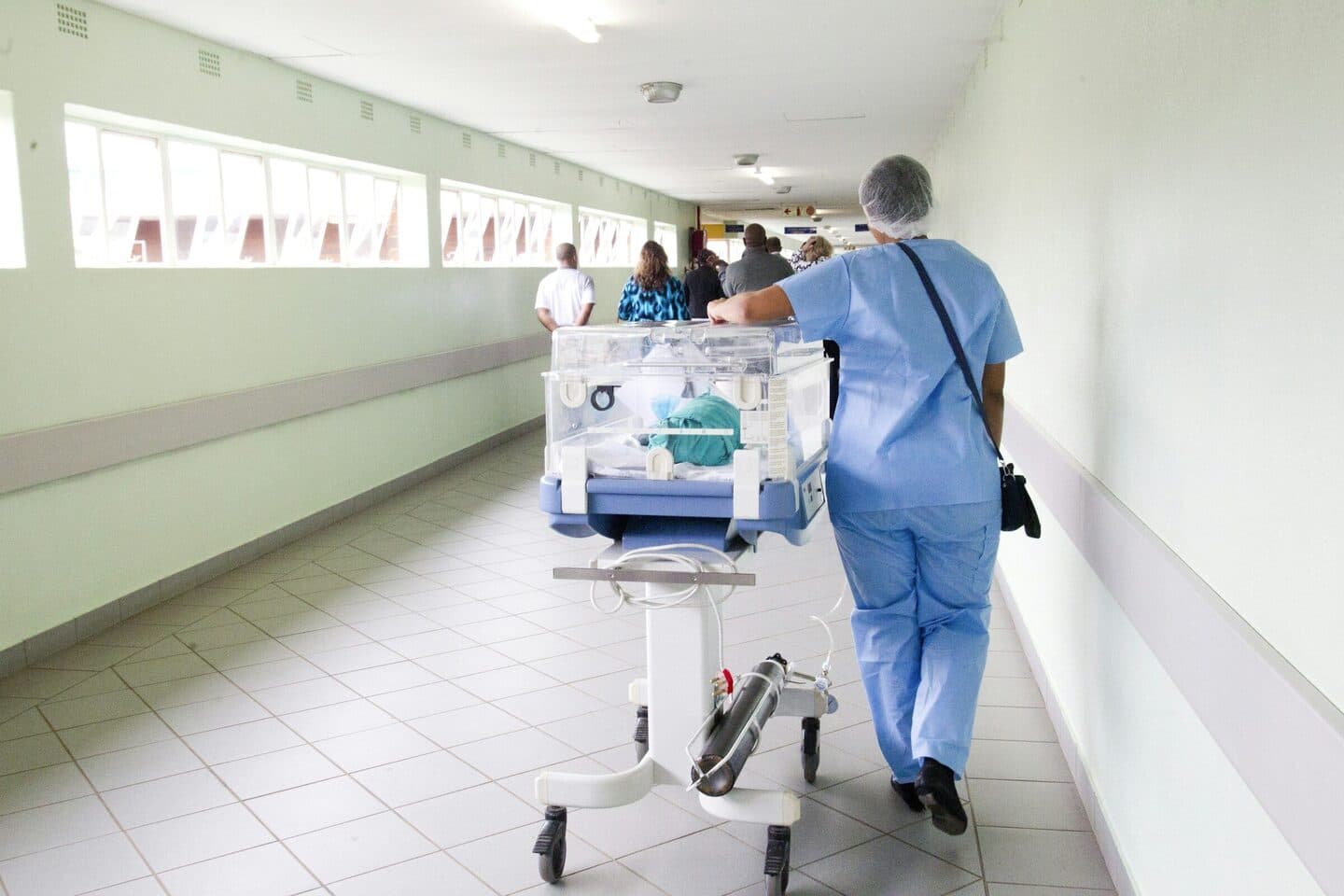 Nurse in blue scrubs pushes an incubator along a hospital corridor